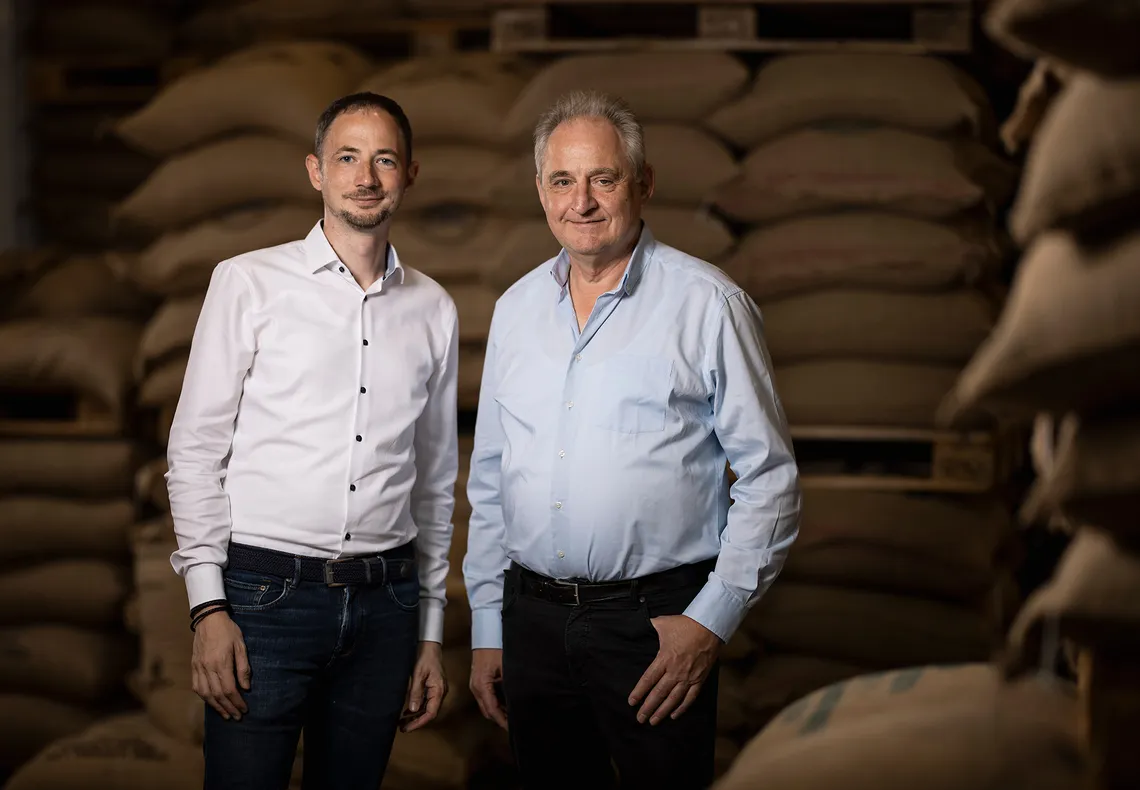 Due uomini della famiglia imprenditoriale Alps Coffee in piedi davanti a sacchi di juta nel magazzino della torrefazione