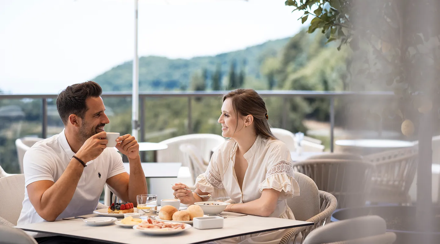 Smiling couple enjoying coffee at breakfast on hotel terrace with mountain view and sun umbrella