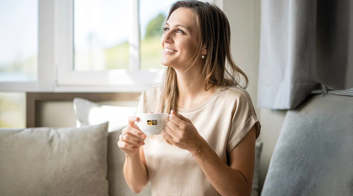 Smiling woman sitting relaxed on sofa, holding Alps Coffee cup and gazing contentedly out the window
