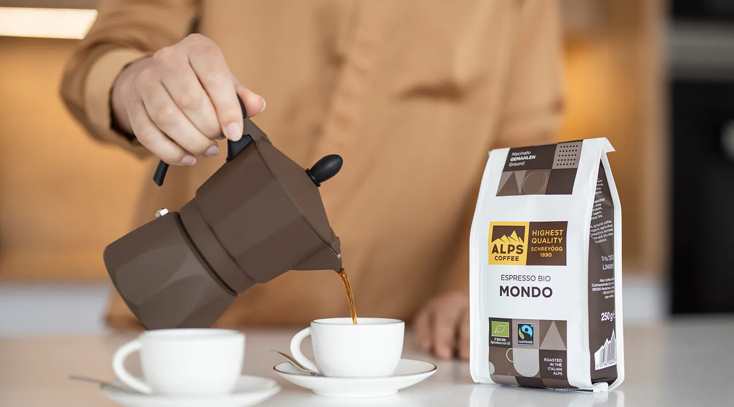Hand pouring espresso from brown moka pot into cup, beside Alps Coffee Espresso Bio Mondo package