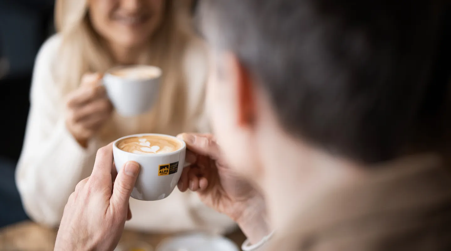 Man holding Alps Coffee cup with latte art, smiling woman with cappuccino in background