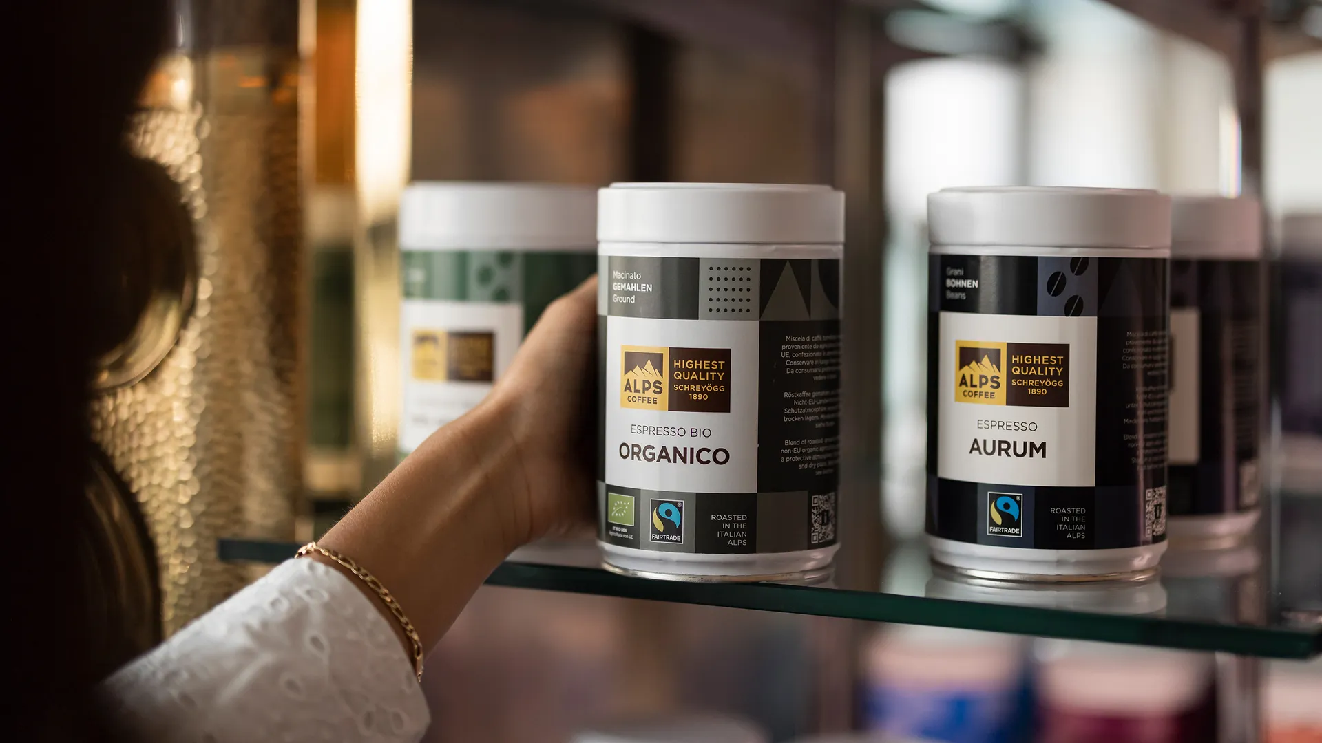 Woman hand reaching for Alps Coffee Espresso Bio Organico tin on glass shelf next to Aurum tin in shop