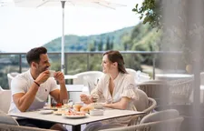 Smiling couple enjoying coffee at breakfast on hotel terrace with mountain view and sun umbrella