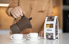 Hand pouring espresso from brown moka pot into cup, beside Alps Coffee Espresso Bio Mondo package