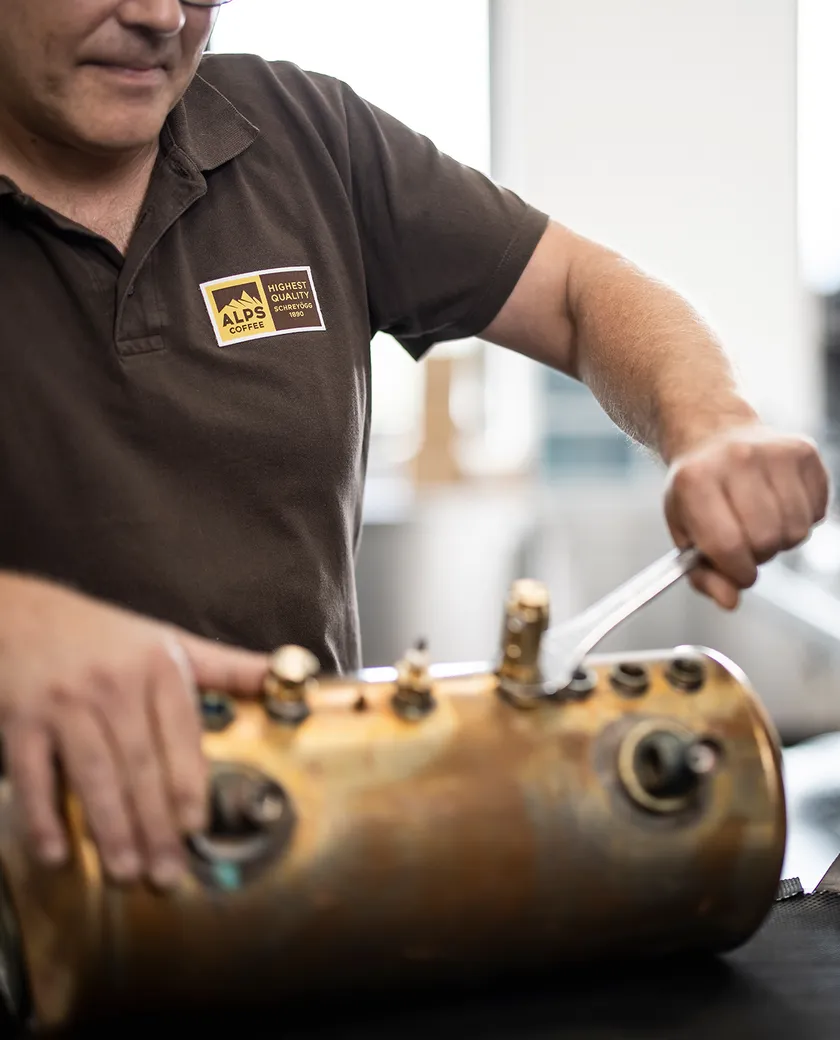 Alps Coffee technician in brown polo maintaining brass brew group of espresso machine with wrench