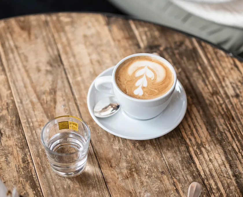 Alps Coffee Cappuccino mit Latte Art Blumenmuster auf rustikalem Holztisch, daneben Alps Coffee Wasserglas