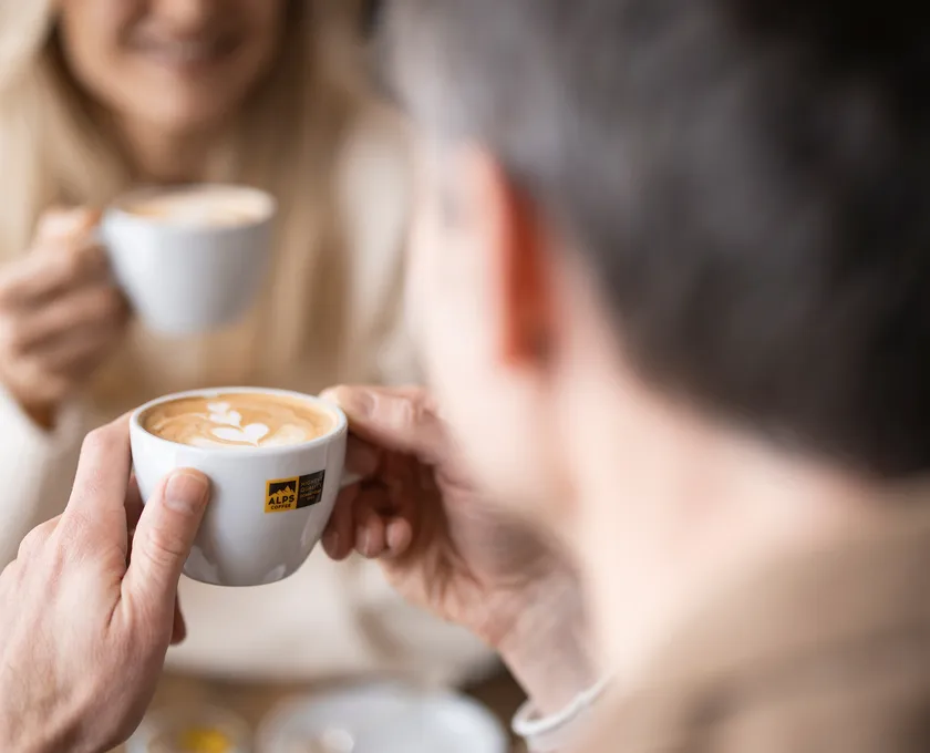 Paar hält Alps Coffee Tassen mit Latte Art — Alps Coffee Logo auf weißer Espressotasse sichtbar
