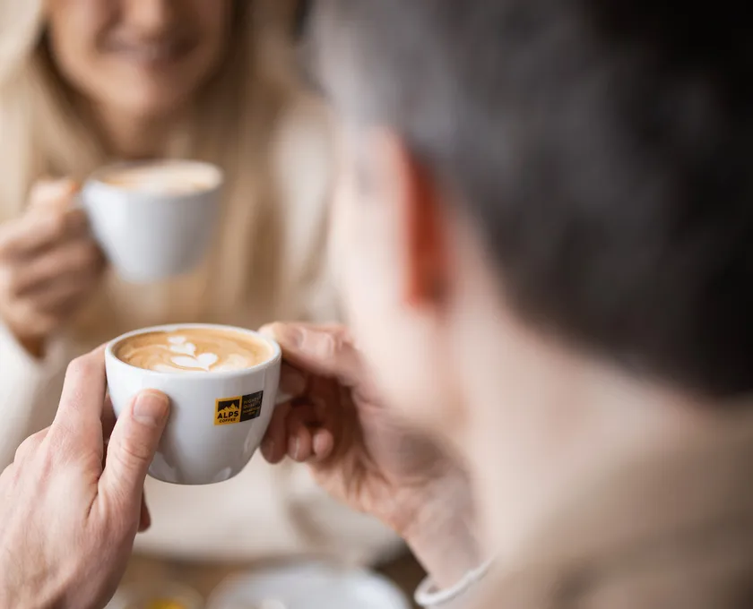Man holding Alps Coffee cup with latte art, smiling woman with cappuccino in background