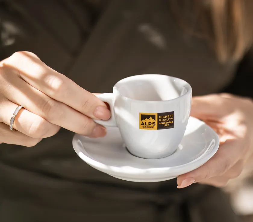 Elegant woman hands holding white Alps Coffee espresso cup on saucer — service moment in store